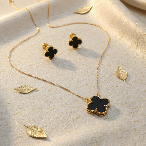 Gold jewelry set with a four-leaf clover pendant necklace, matching stud earrings, and decorative gold leaf accents on a textured cream fabric background.